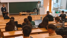 In the lecture hall, you can see the students looking to the front from behind, where two speakers are giving a lecture.