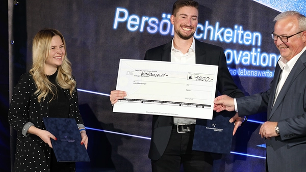 3 people stand side by side on a stage. The man in the middle holds up an oversized check.