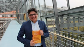 A man in a suit shows an orange book, with a glass THI building in the background.