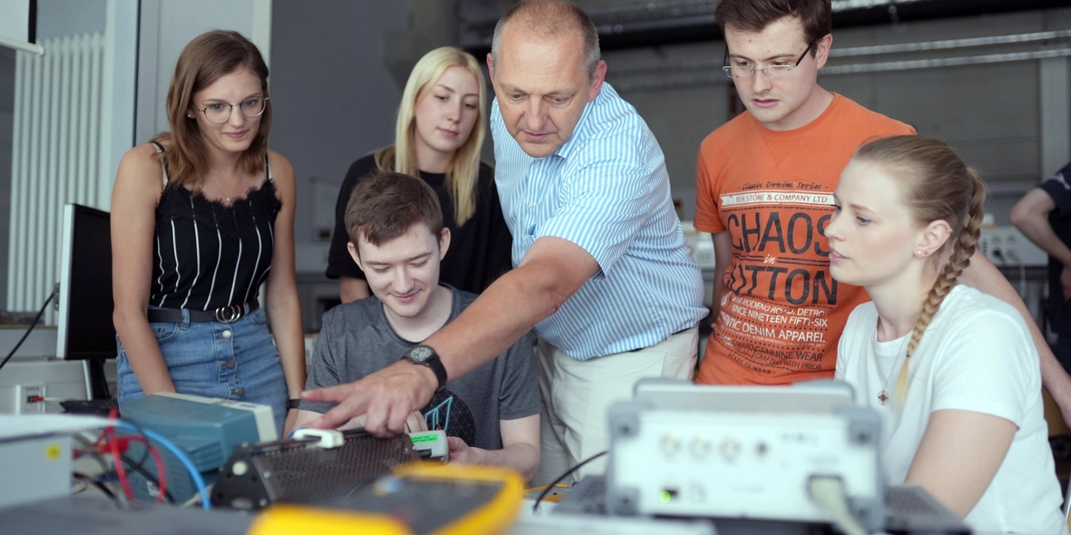 5 Studierende stehen um einen Professor rum. Der Professor deutet auf einen Tisch und erklärt ein Gerät auf diesem Tisch zum Thema Elektrotechnik.