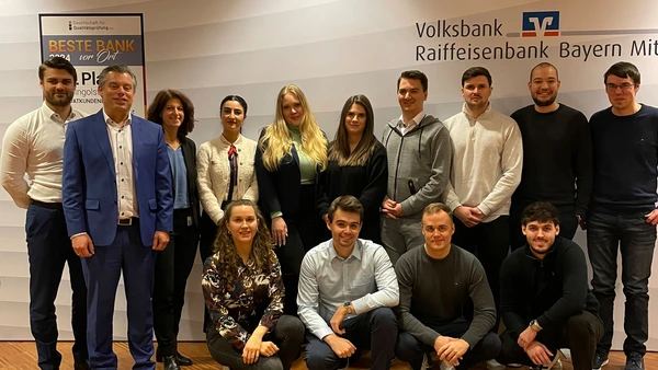 A group photo with 14 people, with the VR-Bank Bayern Mitte logo in the background.