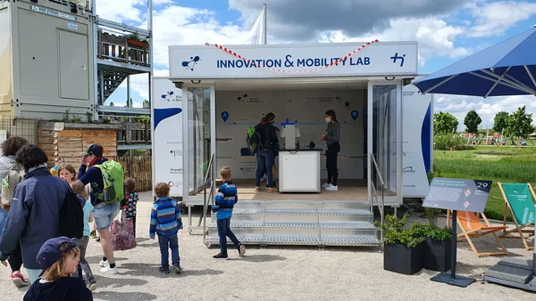 Bild zeigt das Innovation&Mobility Lab von außen mit vorbeigehenden Besuchern und einen mit Wolken bewegten Himmel.