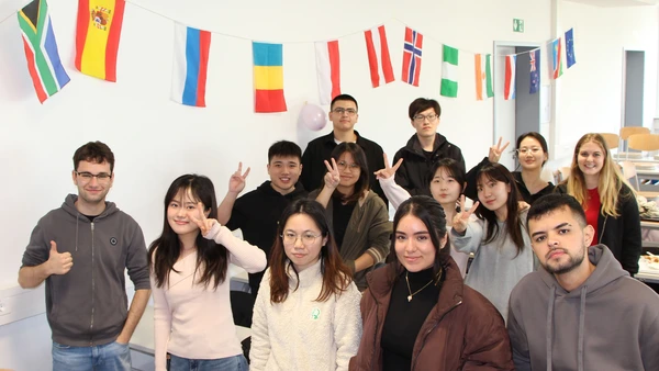 A group of 13 young people smiling into the camera, with various national flags decorated on the wall in the background. 